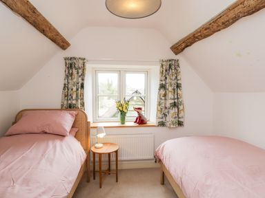 A bedroom with two single beds a small round wooden table with a lamp a window with floral curtains and flowers in a vase at High Cogges Farm Holiday Cottages Witney