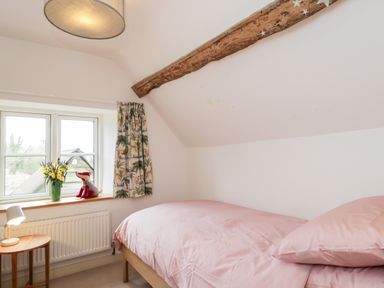 A bedroom with a single bed covered with pink bedding a wooden side table with a lamp a window with curtains and a stuffed toy on the window sill at High Cogges Farm Holiday Cottages Witney