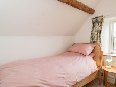 A bedroom with a single bed with pink bedding a wooden nightstand with a lamp a window and patterned curtains at High Cogges Farm Holiday Cottages Witney