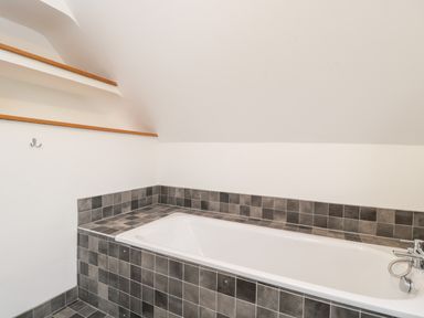 A bathroom with a white bathtub surrounded by gray and black tiles at High Cogges Farm Holiday Cottages in Witney