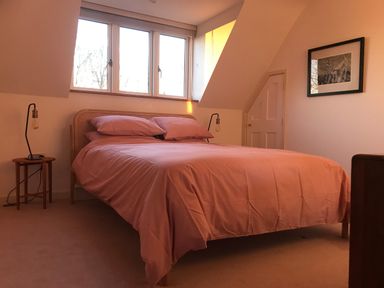 A bedroom with a bed covered in pink bedding two bedside tables with lamps a window a door and a framed picture on the wall at High Cogges Farm Holiday Cottages in Witney