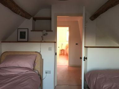 A bedroom with two single beds with pink bedding and wooden beams on the ceiling through an open door a small table with a lamp in an adjoining room at High Cogges Farm Holiday Cottages in Witney