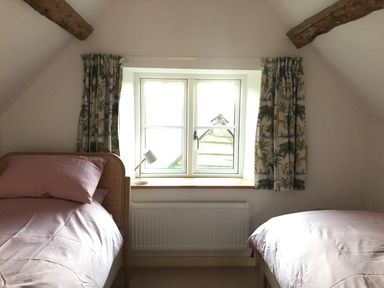 A bedroom with two single beds with pink bedding and a window with patterned curtains at High Cogges Farm Holiday Cottages in Witney