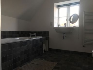 A bathroom with a bathtub surrounded by dark tiles a wall-mounted sink under a window and a rug on the floor at High Cogges Farm Holiday Cottages in Witney