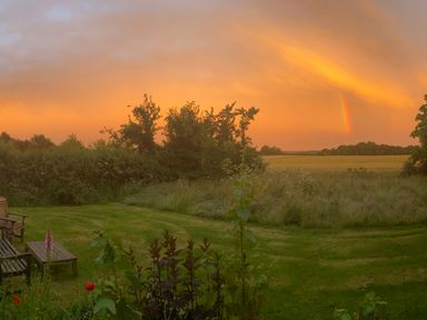 A garden with lawn chairs and table overlooking trees and fields under an orange sky with a rainbow at High Cogges Farm Holiday Cottages in Witney