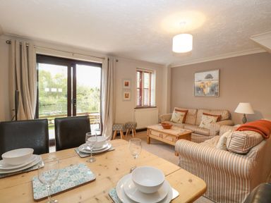 A living room with a wooden dining table set with dishes and glasses a striped sofa set a wooden coffee table and sliding glass doors overlooking a pond at 10 Ruston Reaches in East Ruston near Stalham