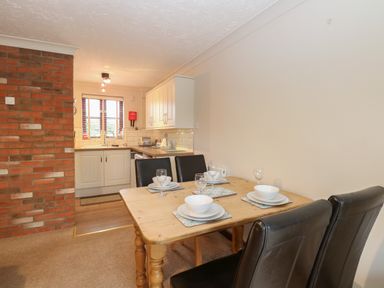 A dining table set with plates and glasses next to a kitchen with white cabinets and a brick wall at 10 Ruston Reaches East Ruston near Stalham