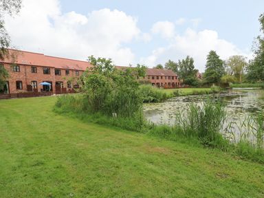 A row of brick houses with brown roofs beside a pond and a grassy area at 10 Ruston Reaches in East Ruston near Stalham