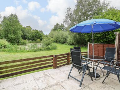 A patio with a glass table and four chairs under a blue umbrella overlooking a fenced grassy area and trees at 10 Ruston Reaches in East Ruston near Stalham