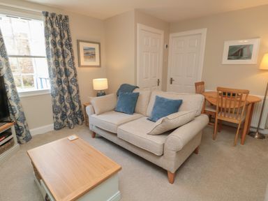 A living room with a beige sofa with blue cushions a wooden coffee table curtains on the window and a small dining table with chairs at Charter's Cottage in Berwick-Upon-Tweed