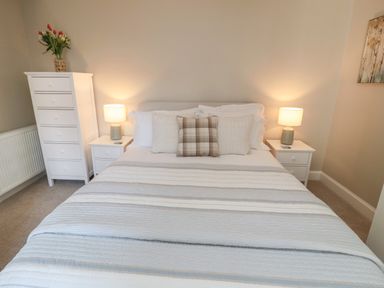 A bedroom with a double bed with pillows and a striped bedspread two bedside tables with lamps a tall white chest of drawers with flowers in a vase on top and a painting on the wall at Charters Cottage in Berwick-Upon-Tweed
