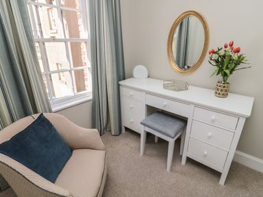 A room with a striped armchair, a white desk with drawers, a mirror, flowers in a vase, and a window with curtains at Charters Cottage in Berwick-Upon-Tweed