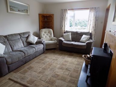 A living room with two sofas, a beige armchair, a wooden corner shelf, a window with curtains, and a fireplace with a stove at Brookfield in St Gennys near Wainhouse Corner