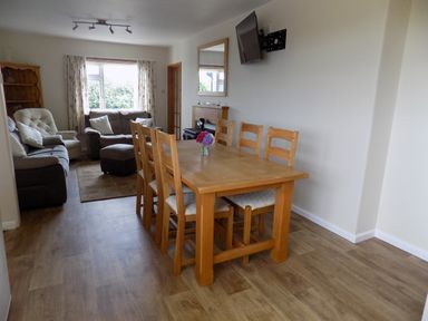 A dining area with a wooden table and six chairs next to a seating area with sofas and an armchair at Brookfield in St Gennys near Wainhouse Corner