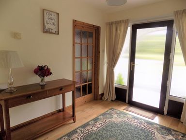 An entrance hall with a wooden table holding a lamp and flowers a glass-paneled wooden door curtains and a rug at Brookfield in St Gennys near Wainhouse Corner