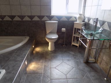 A bathroom with a toilet a glass sink on a metal stand a white cabinet with a pitcher and a bathtub with tile flooring at Brookfield in St Gennys near Wainhouse Corner