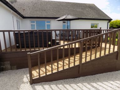 A wooden deck with a ramp railing and outdoor furniture including a table umbrella near a white house at Brookfield St Gennys near Wainhouse Corner