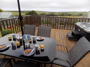 An outdoor wooden deck with a glass table set for breakfast and a hot tub overlooking fields at Brookfield in St Gennys near Wainhouse Corner