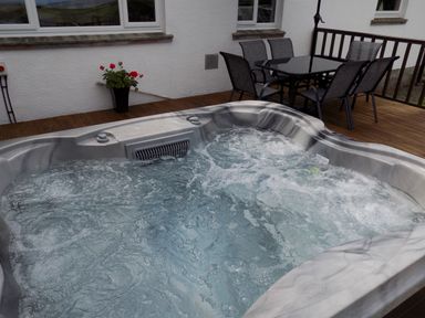 An outdoor hot tub with bubbling water on a wooden deck next to a table with six chairs and a flower pot on a white wall at Brookfield in St Gennys near Wainhouse Corner