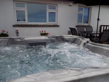 A bubbling hot tub on a patio near a white house with windows and a table with chairs at Brookfield in St Gennys near Wainhouse Corner