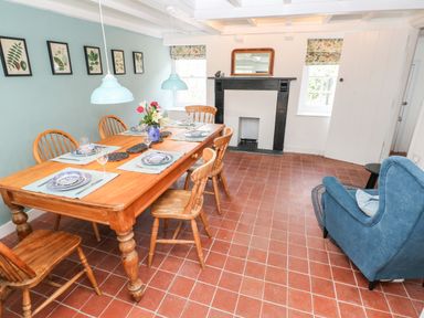 A dining room with a wooden table set for six, wooden chairs, a blue armchair, a fireplace, and botanical prints on the wall at 10 Westgate Hill in Pembroke