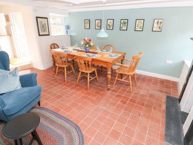 A dining room with a wooden table set with plates and glasses six wooden chairs blue armchair with cushions small round black table and framed botanical prints on blue wall at 10 Westgate Hill in Pembroke
