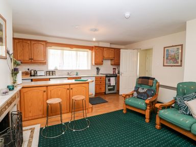 A kitchen with wooden cabinets and stools next to a living area with two green cushioned armchairs at 2 Garnish Court in Glengarriff County Cork