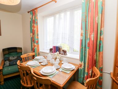 A dining area with a wooden table set with plates and cups four wooden chairs a large window with patterned curtains and a small armchair in the corner at 2 Garnish Court in Glengarriff County Cork