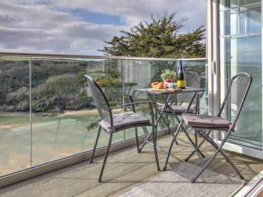 A balcony with a table set for breakfast and three chairs overlooking a beach and wooded area at The Tower 5 Corisande Manor in Newquay