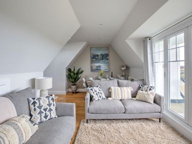 A living room with two grey sofas, a lamp, a plant, and a dining table with chairs by a window at The Tower, 5 Corisande Manor in Newquay