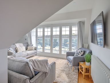 A living room with grey sofas cushions a rug a side table with a plant a wall mounted tv and large glass doors leading to a balcony at The Tower 5 Corisande Manor in Newquay