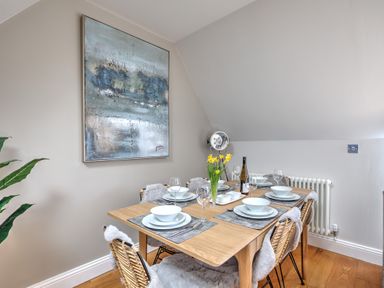 A dining area with a wooden table set with plates bowls and wine glasses surrounded by woven chairs with cushions a painting on the wall and a plant at The Tower 5 Corisande Manor in Newquay