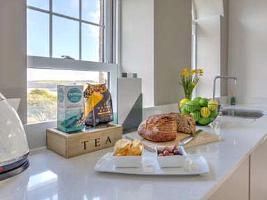A kitchen countertop with bread on a cutting board a plate with chips and olives a basket of lemons and limes and tea boxes at The Tower 5 Corisande Manor in Newquay