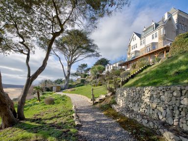 A gravel path next to stone retaining walls with trees grass and a multi story building on a hill at The Tower 5 Corisande Manor in Newquay