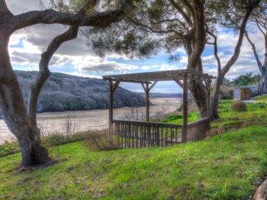 A wooden pergola on a grassy hill overlooking a sandy riverbank with trees and hills in the background at The Tower 5 Corisande Manor in Newquay