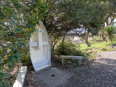 A small white boat propped upright under trees with two wooden benches and a gravel path nearby at The Tower 5 Corisande Manor in Newquay