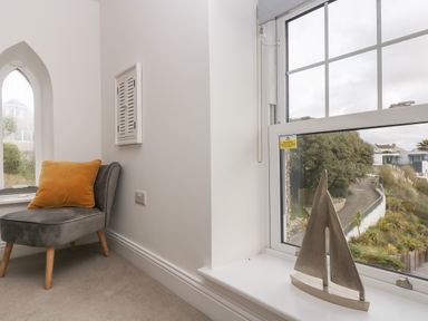 A small corner with a gray chair and orange cushion next to two windows one with a sailboat decoration on the sill at The Tower 5 Corisande Manor in Newquay