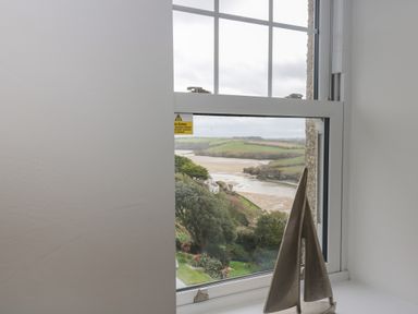 A window with a metal sailboat decoration on the sill overlooking a river and green hills at The Tower 5 Corisande Manor in Newquay