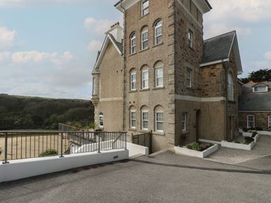 An exterior view of a large stone and stucco building with multiple windows and a metal railing overlooking a sandy area at The Tower 5 Corisande Manor in Newquay