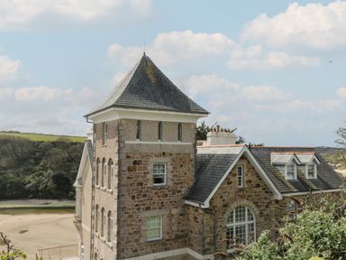 A stone building with a tower and slate roof near a beach and green hills at The Tower 5 Corisande Manor Newquay