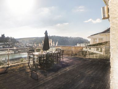 An outdoor wooden deck with a table and six chairs with an umbrella overlooking a waterfront and distant hills at Estuary View in Newton Ferrers