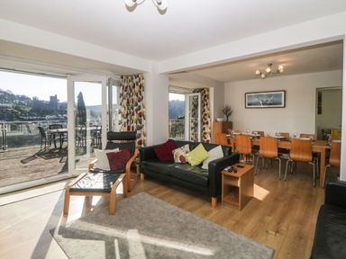 A living room with a black sofa and wooden chair next to large glass doors leading to a balcony and a dining area with a wooden table and chairs in Estuary View in Newton Ferrers