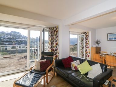 A living room with black leather sofa and chair with cushions a wooden dining table and chairs near large glass doors with a balcony and hillside view at Estuary View in Newton Ferrers
