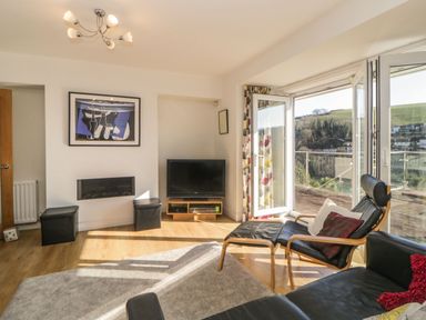 A living room with a black leather chair and ottoman next to an open glass door overlooking hills at Estuary View in Newton Ferrers