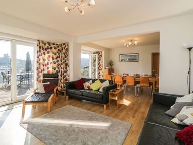 A living room with black leather sofas a wooden armchair a dining table with chairs and glass doors leading to a balcony at Estuary View in Newton Ferrers