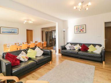 A living room with black sofas and cushions next to a dining area with a wooden table and chairs at Estuary View in Newton Ferrers
