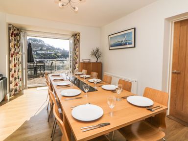 A dining room with a wooden table set with plates and wine glasses and chairs around it and open doors leading to a balcony with outdoor furniture at Estuary View in Newton Ferrers