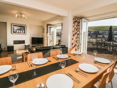A dining area with a wooden table set with plates and glasses next to a living room with black sofas and a television at Estuary View in Newton Ferrers