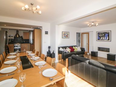 A dining area with a long wooden table set with plates and glasses next to a kitchen and a living room with black sofas and colorful cushions at Estuary View in Newton Ferrers