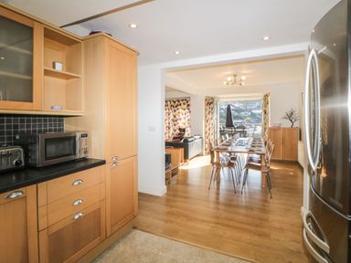A kitchen with wooden cabinets and appliances leading to a dining area with a table and chairs at Estuary View in Newton Ferrers